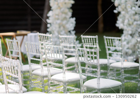 Transparent chairs lining grassy wedding venue, adorned with delicate white flower decorations ceremonial for seating 130950262
