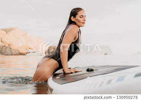 Woman Paddleboard Beach Pose - A woman in a black swimsuit poses on a white paddleboard in shallow water near a sandy beach. 130950278