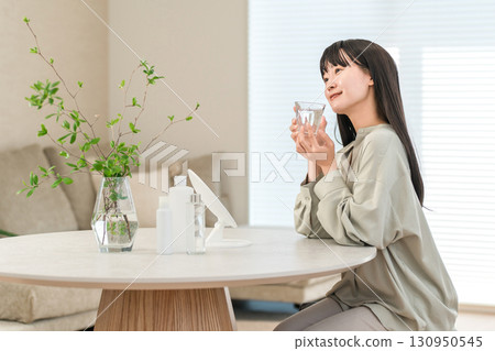 A young Asian, Japanese woman drinking mineral water, water, hot water in a glass in the living room in the morning (detox) 130950545