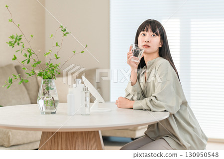 A young Asian, Japanese woman drinking mineral water, water, hot water in a glass in the living room in the morning (detox) 130950548