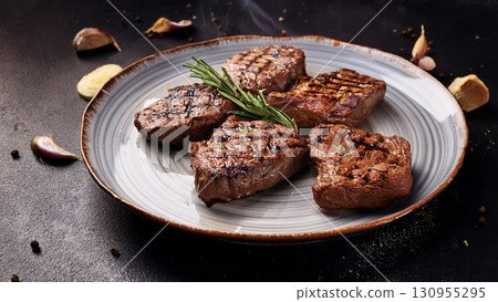 Grilled beef steaks with rosemary, salt, and pepper on wooden board. Juicy barbecue meat with spices. Delicious cooked beef fillet on dark rustic background. 130955295