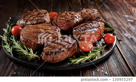 Grilled beef steaks with rosemary, salt, and pepper on wooden board. Juicy barbecue meat with spices. Delicious cooked beef fillet on dark rustic background. 130955297
