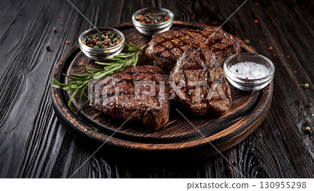 Grilled beef steaks with rosemary, salt, and pepper on wooden board. Juicy barbecue meat with spices. Delicious cooked beef fillet on dark rustic background. 130955298