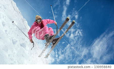 Female skier in pink suit performing jump with skis spread mid-air against snowy slope. Hand made collage 130956640