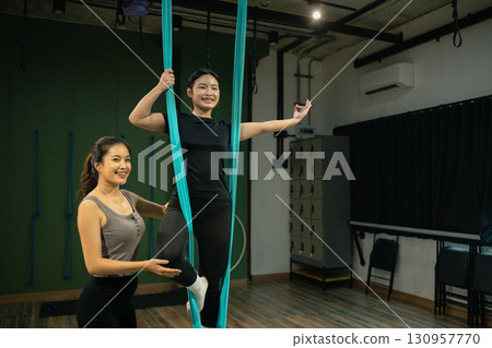 Personal trainer helping woman perform aerial yoga pose, healthy lifestyle and fitness concept 130957770