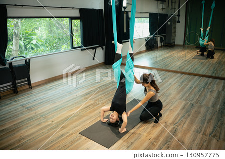 Female trainer assisting young woman in aerial yoga inversion using hammock fabric 130957775