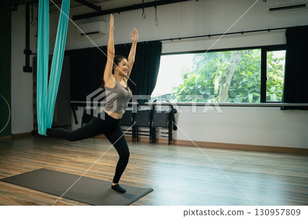 Young female athlete performing aerial yoga exercise with raised arms and extended leg 130957809