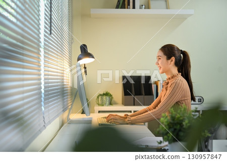 Businesswoman enjoying remote work at organized desk near window blinds Businesswoman enjoying remote work at organized desk near window blinds 130957847
