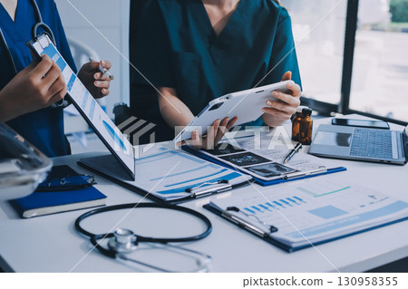 Two doctors and a female nurse meet at a table in the hospital, collaborating on medical tasks using laptops and computers Two doctors and a female nurse meet at a table in the hospital, collaborating on medical tasks using laptops and computers 130958355