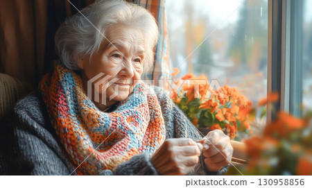 Elderly woman knitting colorful scarf by window with flowers 130958856