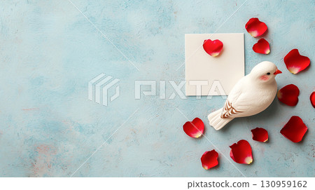 white lovebird figurine surrounded by red rose petals on textured blue background, with blank white card for messages or notes, creating romantic and artistic flat lay 130959162