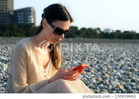 Serious woman browsing smartphone sits on pebble coast in Batumi city Georgia. Serious woman browsing smartphone sits on pebble coast in Batumi city Georgia. 130959668
