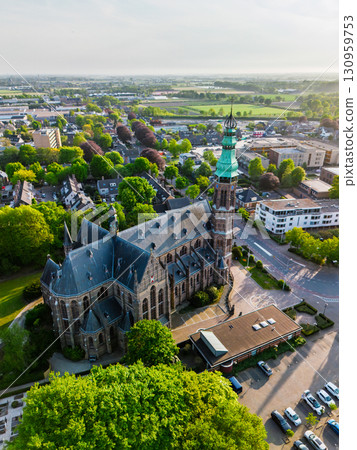 A European town with a tall green-spired Gothic church at its center, surrounded by houses, trees, and streets, glowing warmly in golden hour light. 130959753