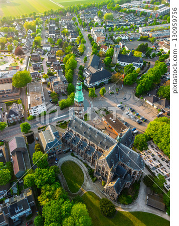 A European town with a tall green-spired Gothic church at its center, surrounded by houses, trees, and streets, glowing warmly in golden hour light. 130959756