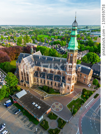A Gothic-style church with a tall green spire and ornate bell tower stands amid gardens, paths, and nearby houses, blending historic elegance with a suburban setting. 130959758