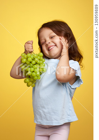 smiling Caucasian little child girl holding bunch of green grapes on yellow background smiling Caucasian little child girl holding bunch of green grapes on yellow background 130959889