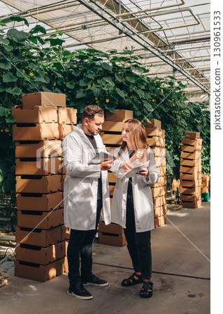 Two agricultural scientists in white lab coats discussing research findings in a greenhouse filled with lush green plants and stacked cardboard boxes, collaboration in sustainable farming practices 130961533