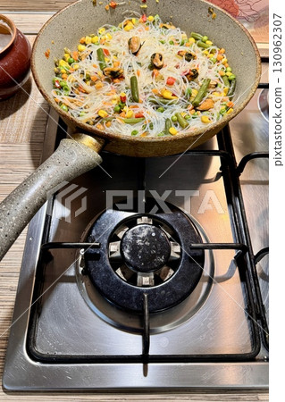 This bright and colorful dish consisting of rice noodles, fresh vegetables and juicy mussels in a pan in a home kitchen is ideal for healthy eating. Close-up. This bright and colorful dish consisting of rice noodles, fresh vegetables and juicy mussels in a pan in a home kitchen is ideal for healthy eating. Close-up. 130962307