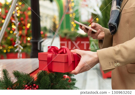 Mans hand holding red gift box as present adorned and using smartphone. Christmas fairy. Xmas shopping. Side view. Close up. 130963202