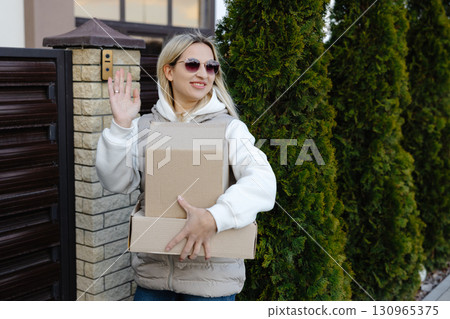 Delivery woman waving while holding cardboard boxes and wearing sunglasses Delivery woman waving while holding cardboard boxes and wearing sunglasses 130965375