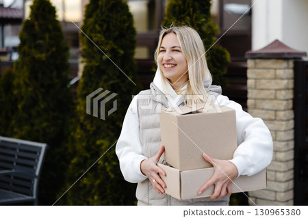 Smiling delivery woman carrying cardboard boxes in front of a house Smiling delivery woman carrying cardboard boxes in front of a house 130965380