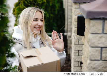 Smiling delivery woman waving while holding a cardboard box Smiling delivery woman waving while holding a cardboard box 130965383