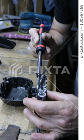 A repairman's hands with a small soldering iron while restoring a screwdriver battery, working with a hand soldering iron on a workbench surface while repairing a screwdriver battery 130967869