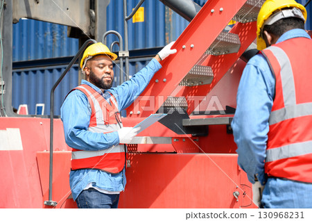 Two African American logistic workers wearing reflective vests and white helmets talk about logistics operations at shipping container yard. Transportation import and export logistic industry concept. Two African American logistic workers wearing reflective vests and white helmets talk about logistics operations at shipping container yard. Transportation import and export logistic industry concept. 130968231