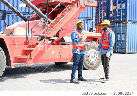Two African American logistic workers wearing reflective vests and white helmets talk about logistics operations at shipping container yard. Transportation import and export logistic industry concept. 130968234