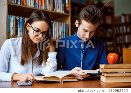 Cheerful students working together in the library, looking through books, looking for information, helping each other in studying Cheerful students working together in the library, looking through books, looking for information, helping each other in studying 130968323