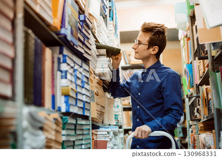 Male student looking for necessary book on numerous shelves in the library. Happy student finally finds book with important information 130968331