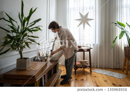 Calm woman reaching over table with cleaning duster tool to wipe surfaces and maintain home hygiene 130968938