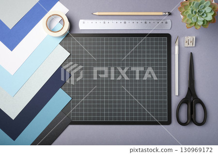 Flat lay of craft supplies including colored paper, cutting mat, scissors, ruler, pencil, masking tape, utility knife, sharpener on a gray background. Copyspace 130969172