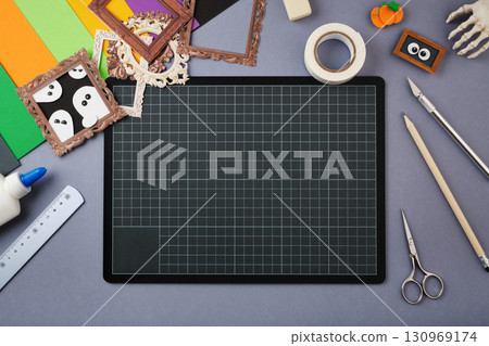 Flat lay of Halloween-themed craft supplies including colored paper, small frames, glue, scissors, ruler, tape, pencils, cutting knife, and spooky decorations around a black cutting mat. Flat lay of Halloween-themed craft supplies including colored paper, small frames, glue, scissors, ruler, tape, pencils, cutting knife, and spooky decorations around a black cutting mat. 130969174