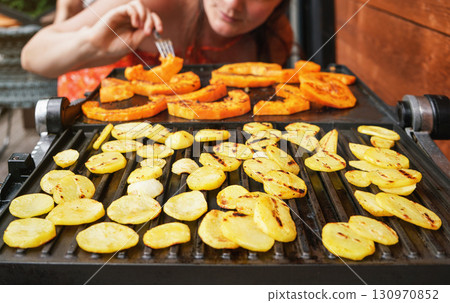 Potato chips and butternut squash pieces grilled on electric grill, focus on bright potatoes circles, blurred young woman in background 130970852