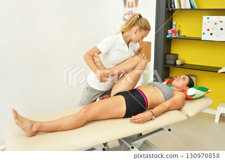 Young female physiotherapist exercising with her patient laying on massage table, flexing her knee muscles 130970858