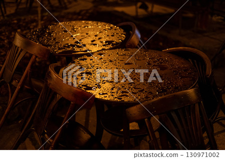 Two rain-speckled cafe tables with wooden chairs sit quietly on a paved patio, bathed in warm light, evoking a moody, tranquil post-rain atmosphere. 130971002
