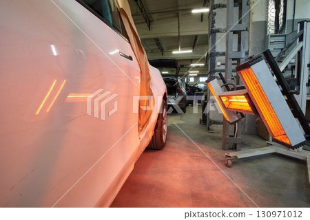 A white car with side damage stands in a repair shop, illuminated by red infrared heat lamps for paint curing, surrounded by tools and other vehicles in a busy garage setting. 130971012
