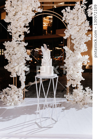 Elegant white wedding cake with sugar floral decor on modern stand at luxury reception 130972109
