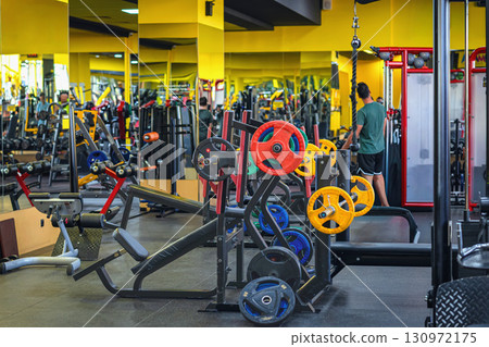 Modern gym interior with colorful weight plates, workout benches, exercise machines, bright yellow fitness space. Concept of gym training, fitness lifestyle, strength exercise in gym, active lifestyle Modern gym interior with colorful weight plates, workout benches, exercise machines, bright yellow fitness space. Concept of gym training, fitness lifestyle, strength exercise in gym, active lifestyle 130972175