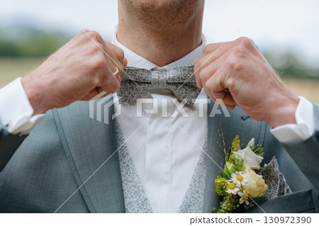 A person adjusts their bowtie on a wedding day, reflecting attention to detail and preparation. The image showcases a suit with a floral pin, capturing an elegant and joyful moment. A person adjusts their bowtie on a wedding day, reflecting attention to detail and preparation. The image showcases a suit with a floral pin, capturing an elegant and joyful moment. 130972390