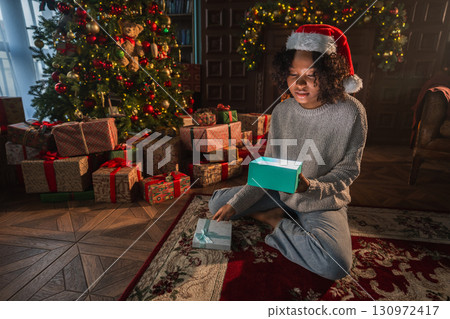 Merry Christmas. African American woman unpacking open up gift box near Christmas tree. Girl in living room with Christmas tree and fireplace opening gift box with surprise face. Christmas eve at home Merry Christmas. African American woman unpacking open up gift box near Christmas tree. Girl in living room with Christmas tree and fireplace opening gift box with surprise face. Christmas eve at home 130972417