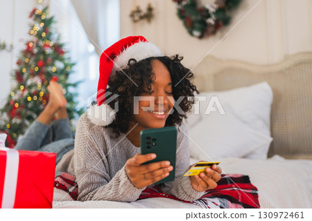 African American woman shopping online holding smartphone paying with gold credit card for Christmas gifts. Girl buying on Internet enter credit card details enjoying Christmas winter holidays at home African American woman shopping online holding smartphone paying with gold credit card for Christmas gifts. Girl buying on Internet enter credit card details enjoying Christmas winter holidays at home 130972461