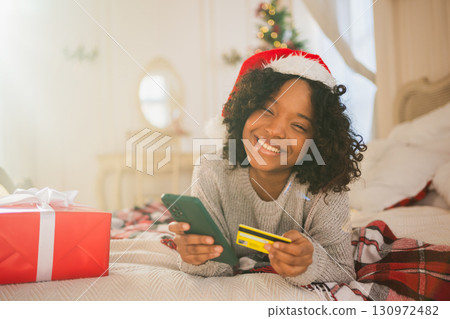 African American woman shopping online holding smartphone paying with gold credit card for Christmas gifts. Girl buying on Internet enter credit card details enjoying Christmas winter holidays at home 130972482