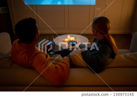 Romantic couple drinking wine from glasses watching movies together on sofa at home on date night Romantic couple drinking wine from glasses watching movies together on sofa at home on date night 130973148