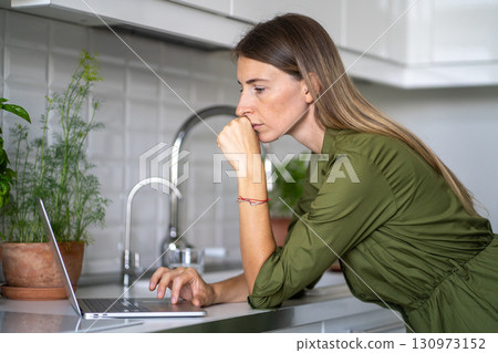 Serious woman working on laptop at kitchen table during break from household chores at home Serious woman working on laptop at kitchen table during break from household chores at home 130973152