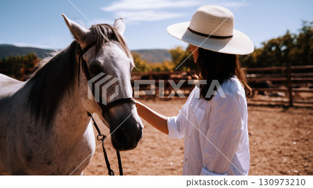 Woman in white shirt and hat gently touching a gray horse on a sunny day at the paddock with mountains in the background 130973210