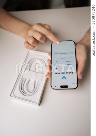 Ufa, Russia - 17 August 2025. Female customer setting up brand new iPhone 16 pro by connecting to wi-fi network after unpacking, with charging cable visible in box, close-up. 130973246