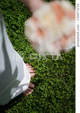 Barefoot bride standing on green grass holding a blurred bouquet on a sunny wedding day 130973254