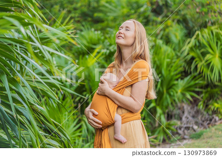 Mother holding her little baby in a yellow sling in the park. Warm and loving family moment. Babywearing, parenting, and mother-child bonding concept 130973869